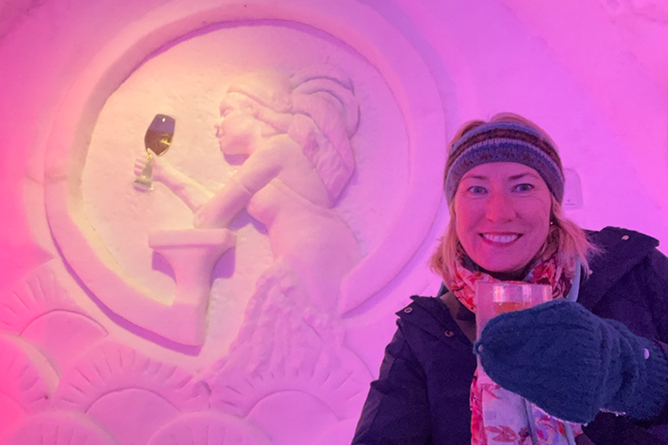 A blonde woman in winter clothing with a drink in front of an ice sculpture