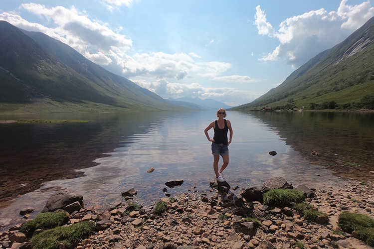 A blonde woman in sunglasses and shorts standing at the edge of reftlective loch on a sunny day