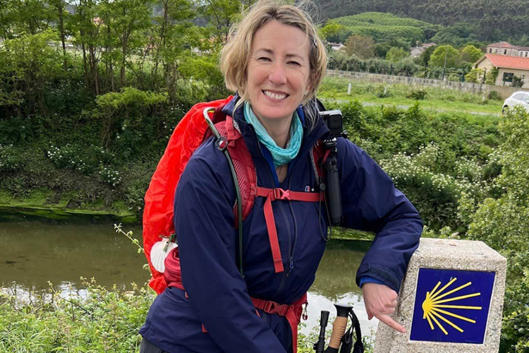 A blonde woman in hiking gear smiling pointing to a blue and yellow sign