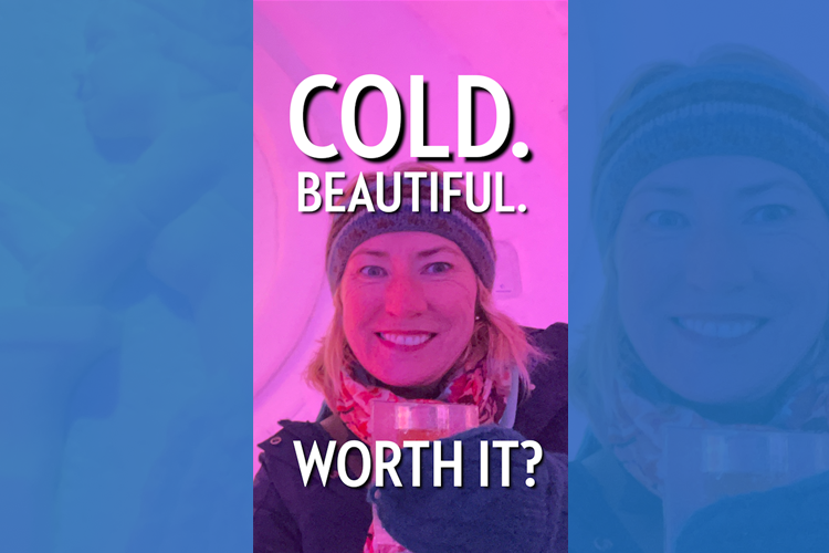 A blonde woman in a wooly hat standing in front of a pink lit wall in an ice hotel holding a cold drink in gloves