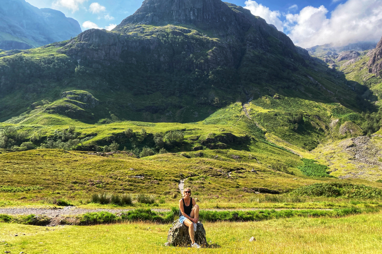 A Woman in a black vest and sunglasses standing beside a calm loch in Scotland with a mountainous backdrop sitting on a rock