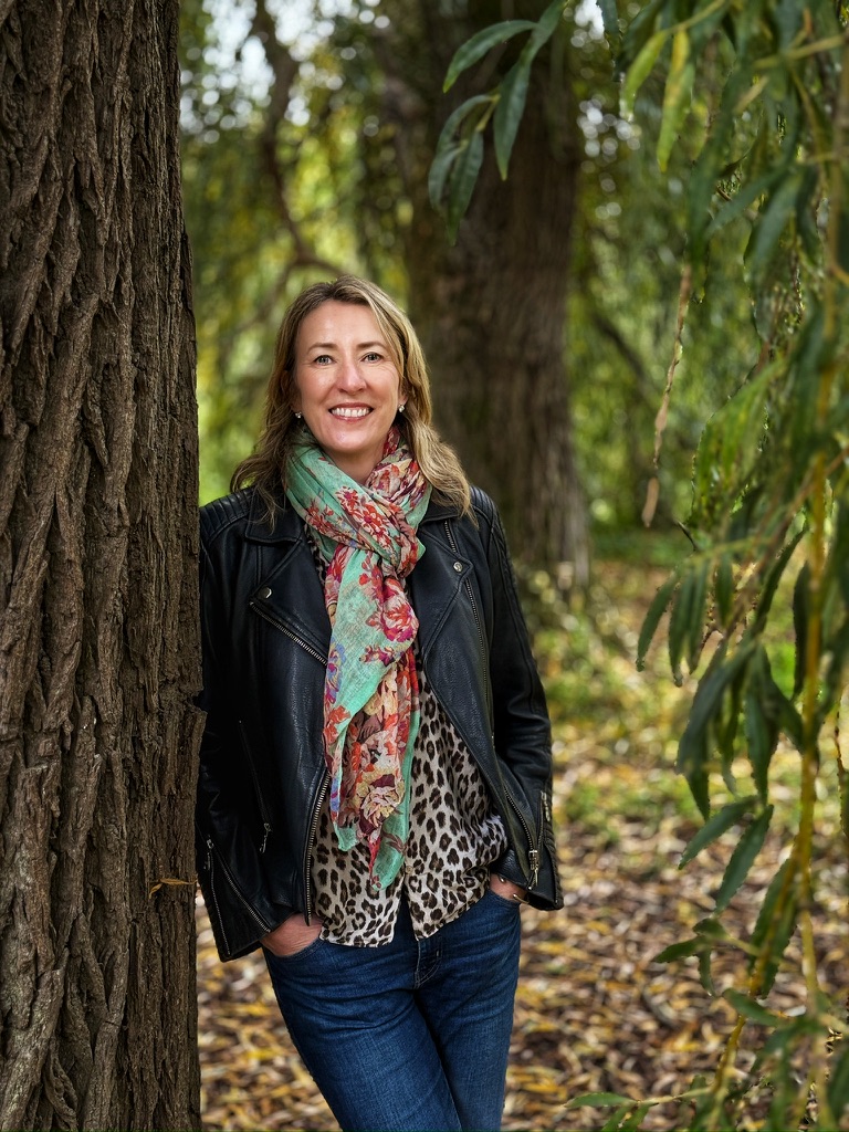 A blonde woman smiling in a patterned scarf leaning up a tree