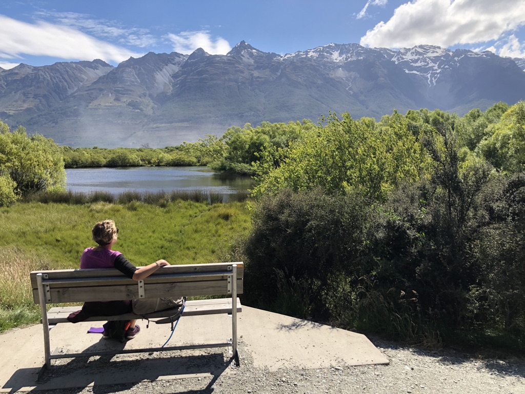 A woman sitting on bench in the sun facing a mountain scene