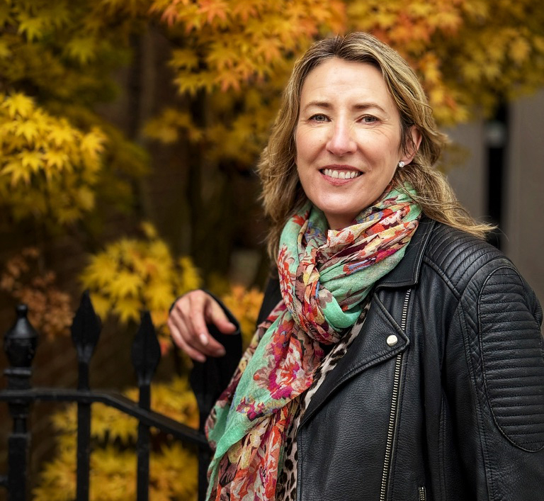 A blonde woman in a colourful scarf standing by a tree